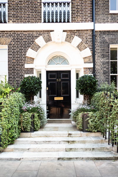 black door, topiary and foliage on either side of the path leading up to it, four broad stone steps, archway above the door with alternating brickwork and white plaster