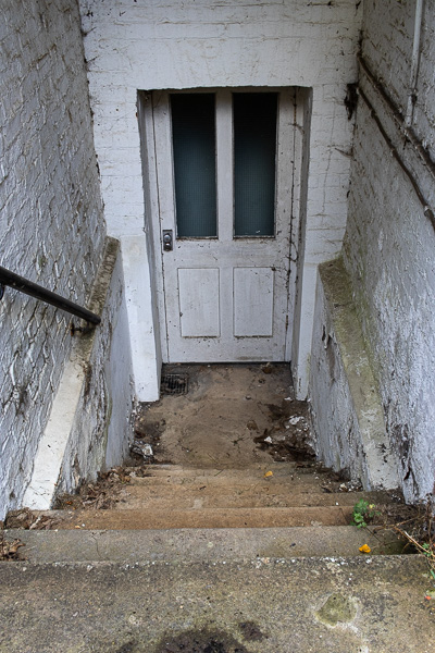 a set of stone steps leading down to an old white painted wooden door as if to a cellar.  The brick walls on either side of the steps are painted white.  There is a black metal handrail on the left hand wall and litter and detritus in the stairwell.