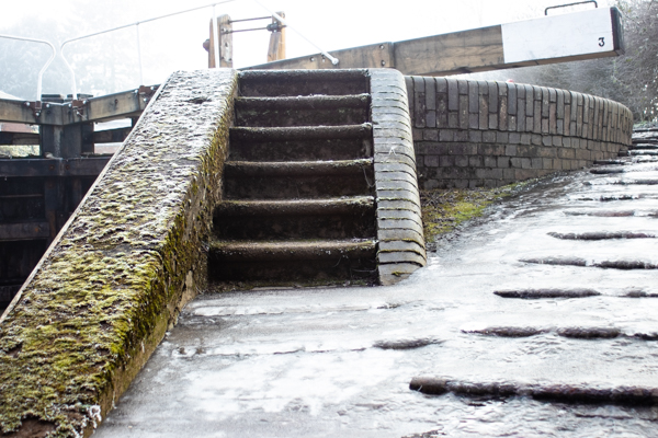 Colour photograph with a set of stone steps leading up to a black and white lock gate. There is a circular wall and a ramp which is covered with ice.