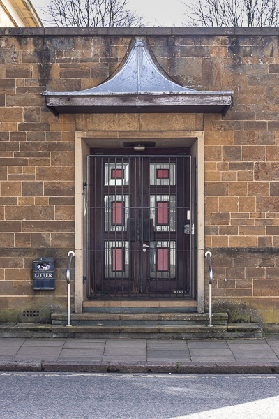 a set of three stone steps leading up towards a double door.  The door is brown with rectangular insets which look like reflective glass with a red rectangle inside those.  There is a barred gate across the doors.  The walls on either side are local yellow stone, and there is a litter bin on the wall.  Above the door is a wooden beam  with a triangular construction above that.
