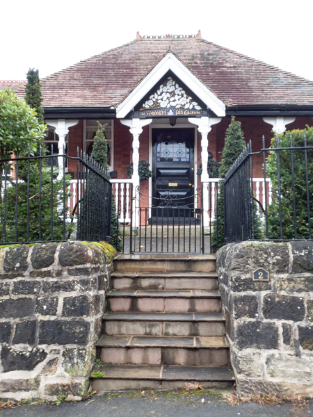 Colour photograph with a set of stone steps leading to a black iron gate and fencing.  Behind is a brick house, with a black door and white columns holding up a porch with a triangular section above the door into the roof area decorated with a white flower design 