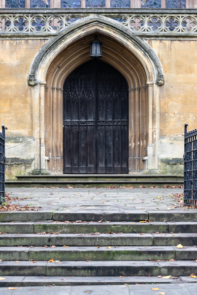 Colour photograph with a set of stone steps and a pair of open black gates.  Behind is a large black door with a pointed stone arch over it. The church is made of yellow coloured stone, the steps are more grey in colour.