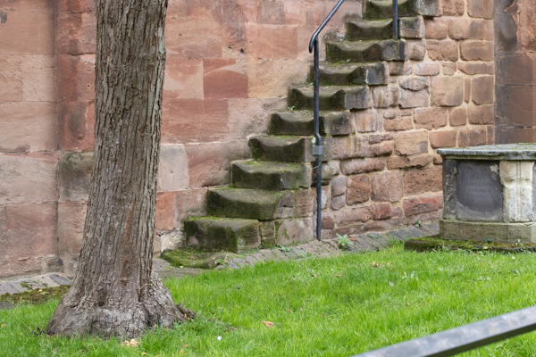 Colour photograph with a set of stone steps in front of a red stone wall of a church.  At the left is a tree trunk and on the right of the picture is a stone tomb both of which are in some green grass.