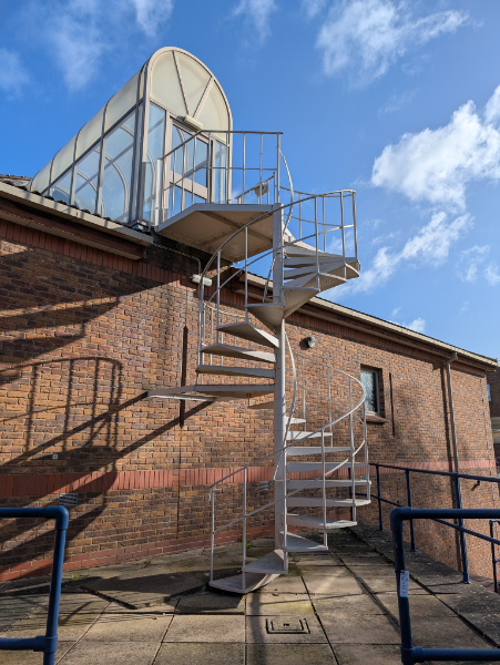 a white spiral staircase up the outside of a brick wall leading to a door in a glass entrance on the roof.