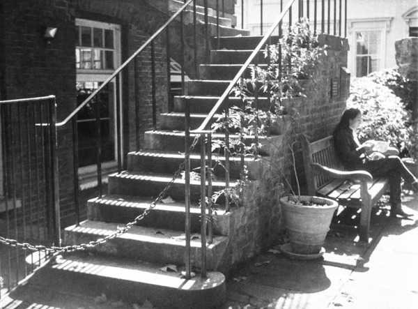Black and white photograph with a set of stone steps from bottom left to top centre, with a bench to one side and a yong lady reading sitting on the bench