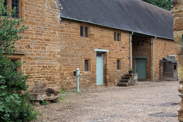 an old brick farmhouse building with leaded windows and a pale green water pump and pale green door. A mounting block with four steps is near to  large entranceway.  The floor is cobbled