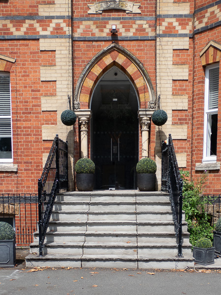 a set of stone steps leading up to a glass door that looks black from the outsidewith a pointed arch above and a slim stone column on each side.  There is a black railing on either side of the steps and a pot with a round ball shaped box shrub in it on either side of the door.  A hanging basket on each side of the door is round.  The door is in a red brick wall with light stone patterning.