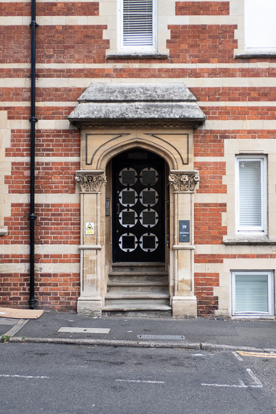 a set of stone steps leading up to a black door with eight white circles in a two by four grid each with a black square inside.  The doorway is surrounded by a brick wall which has stone stripes across and several windows in it.