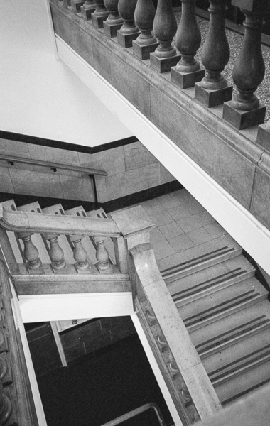 a black and white film photograph of a set of stone indoor steps from left to right with a ninety degree bend.  There are large stone balustrades along the side and across the top right hand corner of the picture