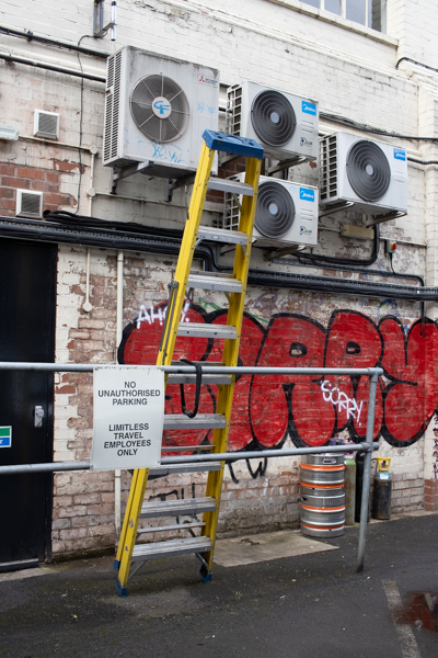 set of metal stepladders leaning against a railing, a white brick wall behind has air conditioning units and red graffiti, there is a beer keg on the floor and a printed sign saying no unauthorised parking Limitless Travel Employees Only.  