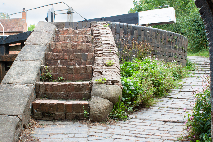 colour image of a set of steps leading up to a black and white lock gate with a number 2.  Sweeping wall on the right of the photo.  A bridge arch on the right of the photo